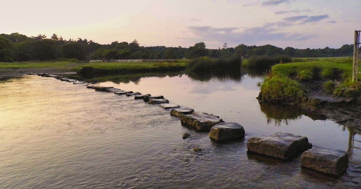 A line of stepping stones crosses a calm river at dusk toward the far bank.