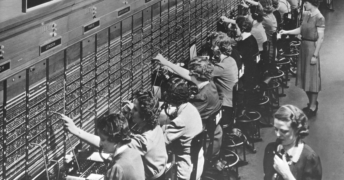 Telephone operators working a crowded Bell System switchboard in 1943, plugging cords into a wall of jacks.