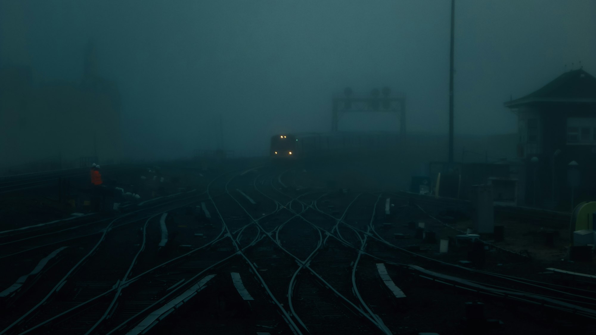 Dozens of railway tracks branch and cross at switches beneath thick blue-hour fog as a lone train's headlights glow in the distance.