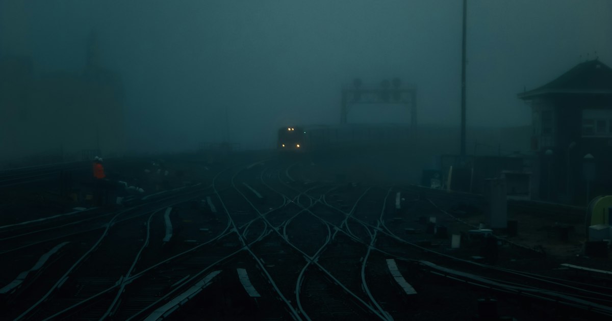 Many railway tracks cross and branch at switches in a foggy, dark blue rail yard as a train approaches in the distance.